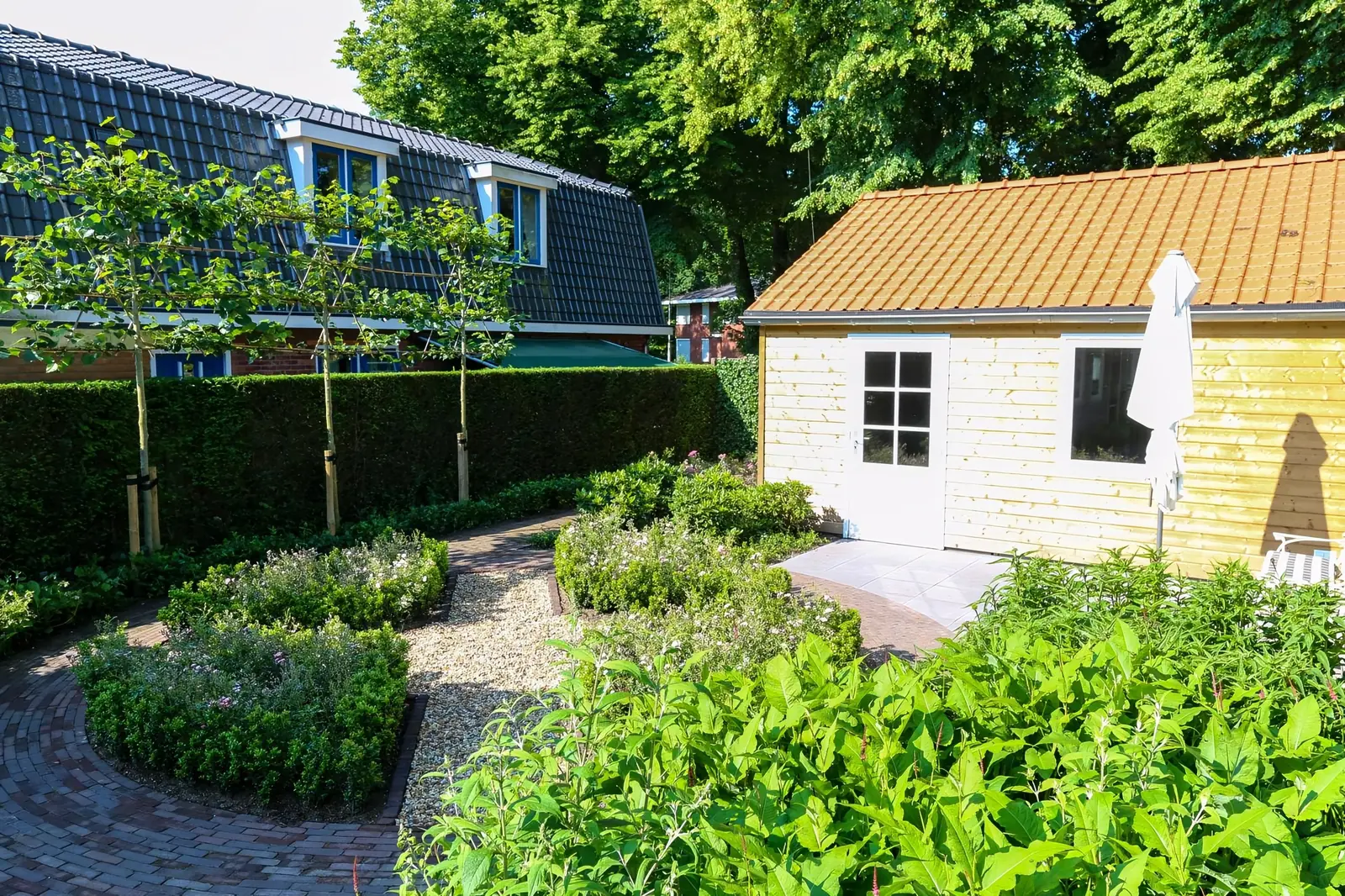 Ronde vakken met lage hagen en klinkerpaden voor een houten tuinhuis met pannendak in Wolfheze